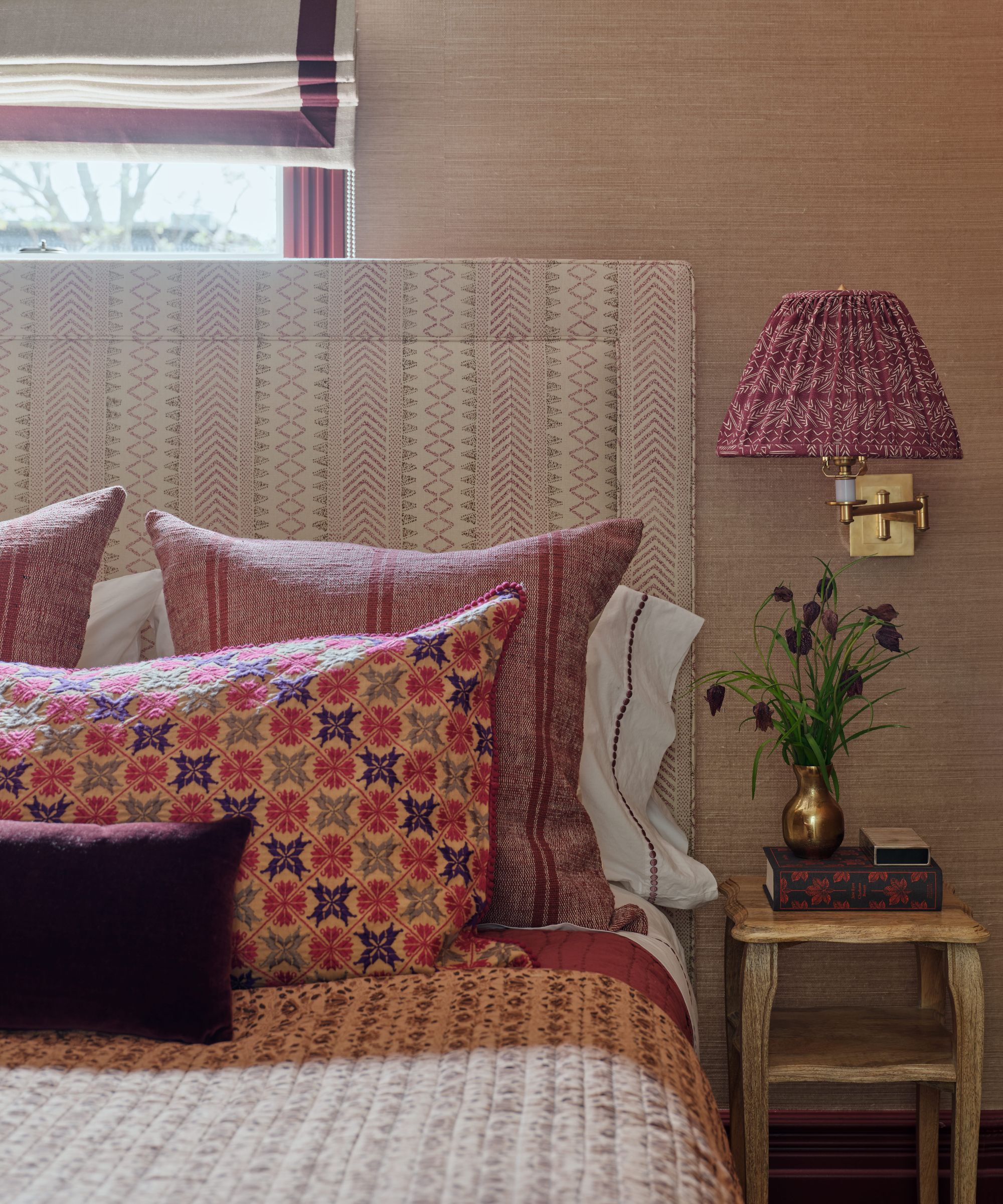 a grasscloth wrapped bedroom with a bed in front of a window with pink and aubergine tones of fabric on headboard, bed linens and small wall sconce