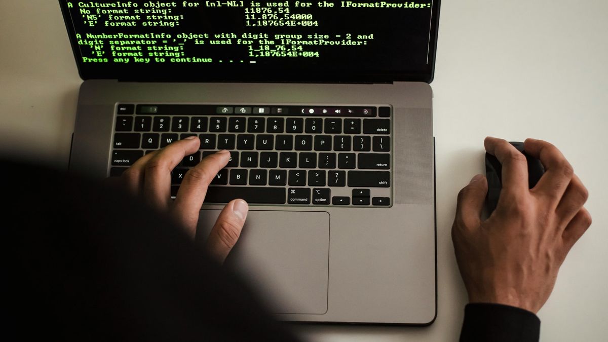 A hacker typing on a MacBook laptop with code on the screen.