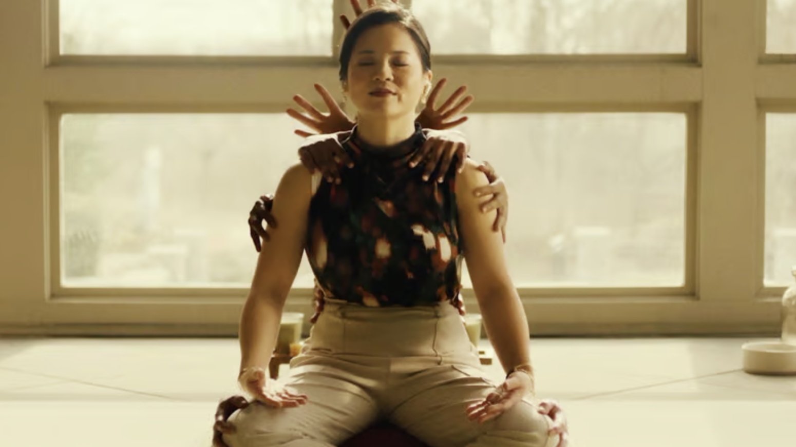 A still from the Hulu movie Control Freak of Kelly Marie Tran sitting cross-legged as hands appear behind her