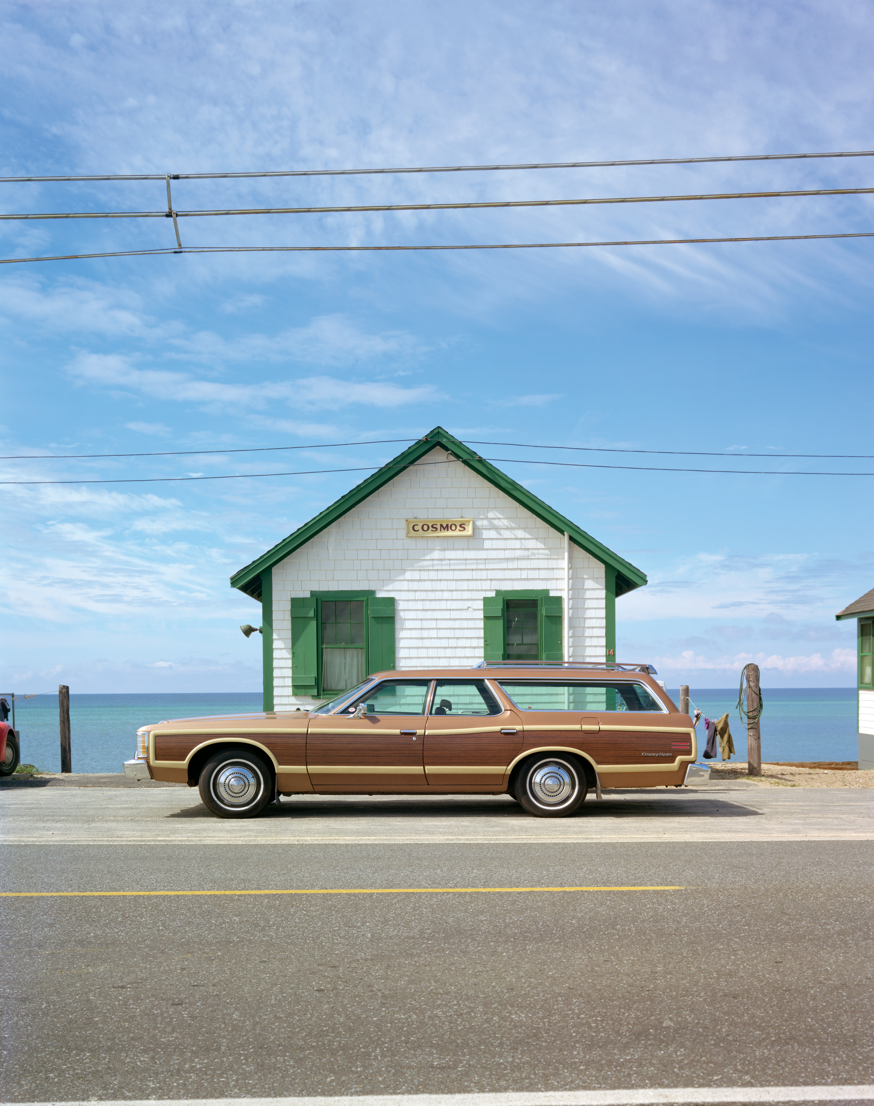 A wood-panelled station wagon is parked outside a small white cottage with green trim and a Cosmos sign, the sea stretching out behind under a pale blue sky.
