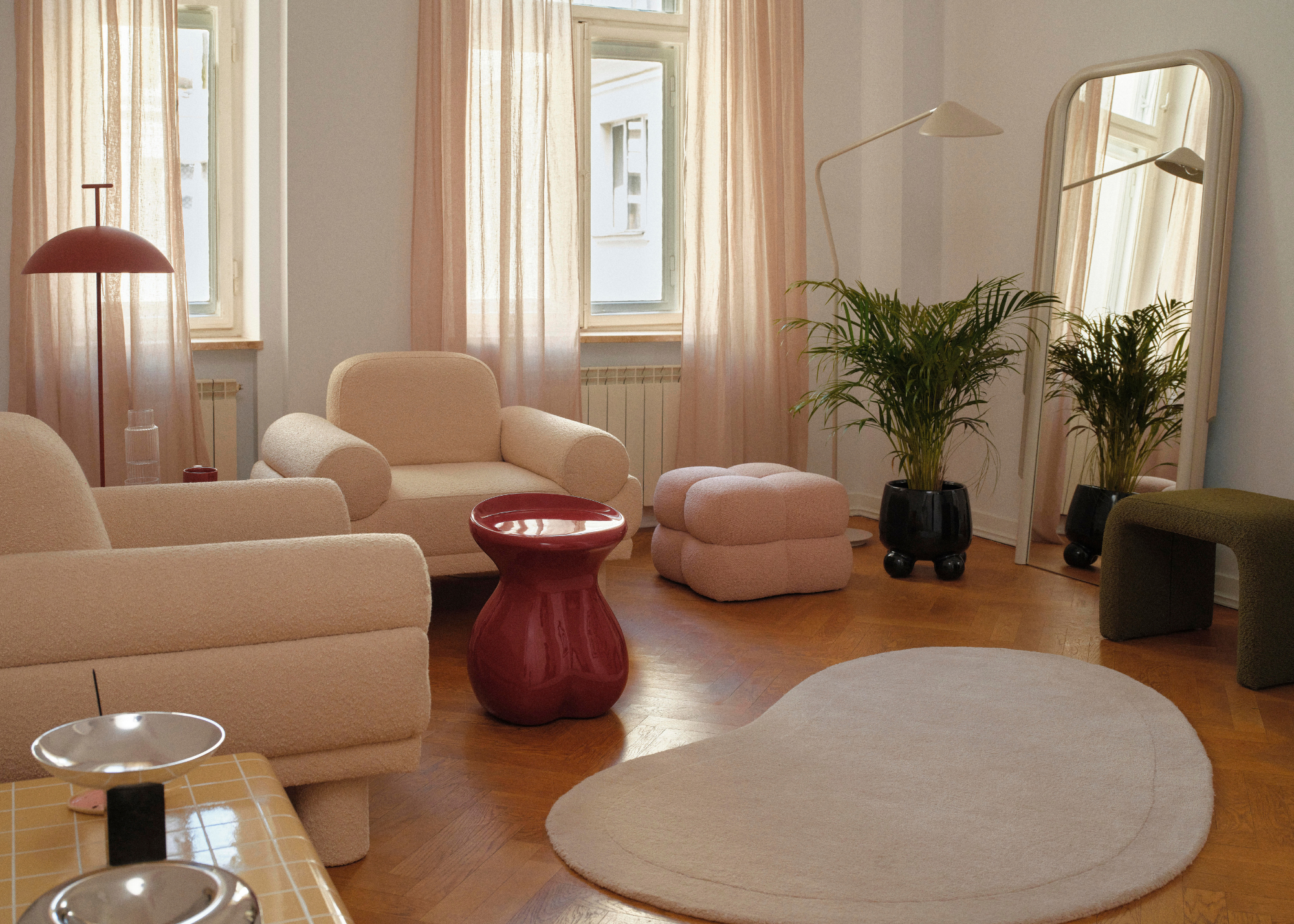 A bright living room with wood flooring, a curved off white rug, a red side table, a pink ottoman, white armchairs, a potted houseplant, and a leaning mirror