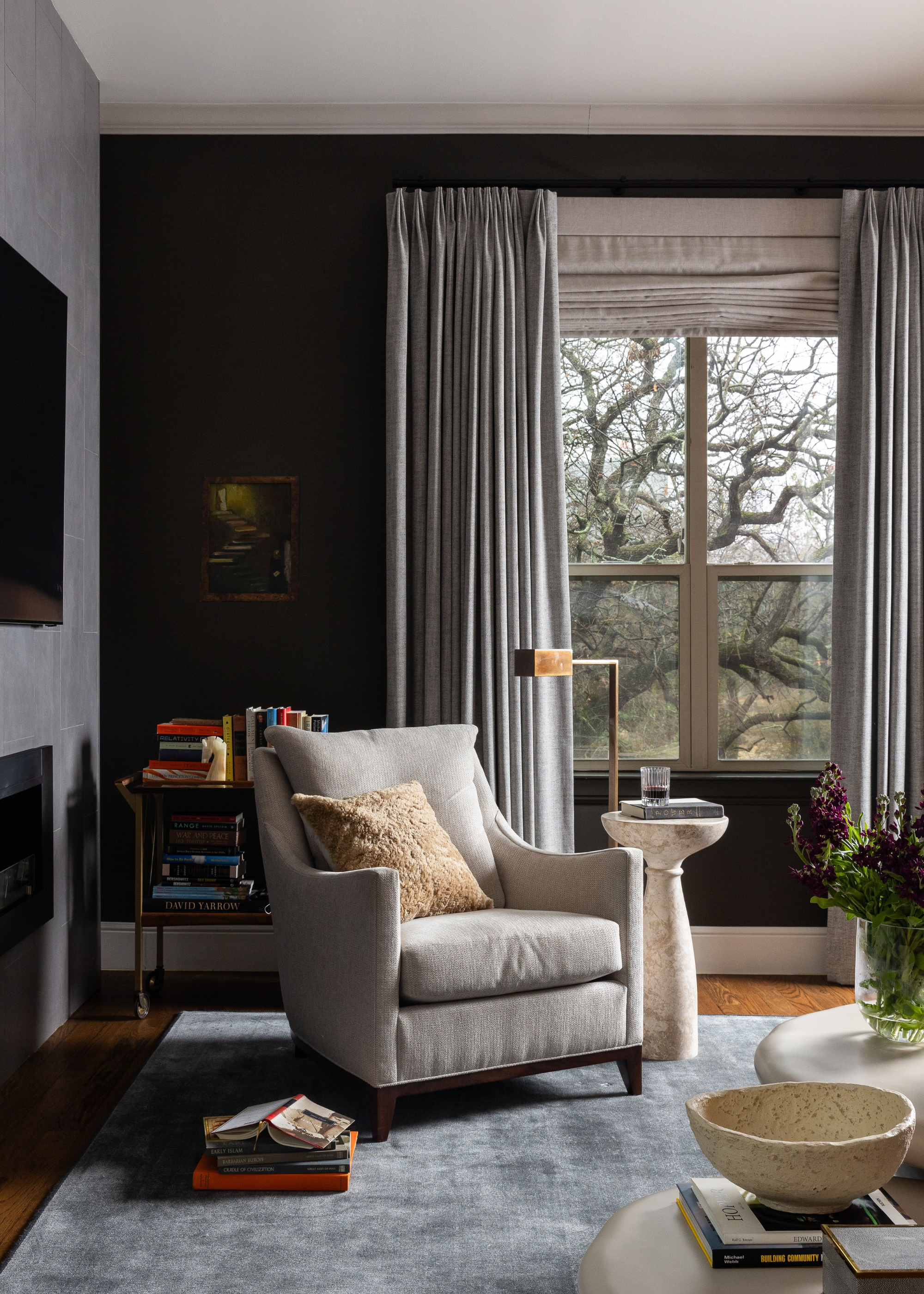 A dark living room with a vintage bar cart holding books beside a gray sofa chair, a standing gold lamp, and a stone side table