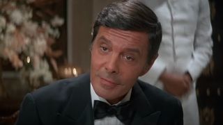 Louis Jourdan smiles while sitting at the table in a tuxedo in Octopussy.