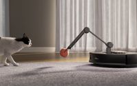 A robot vacuum with an arm holding a ball while a cat watches