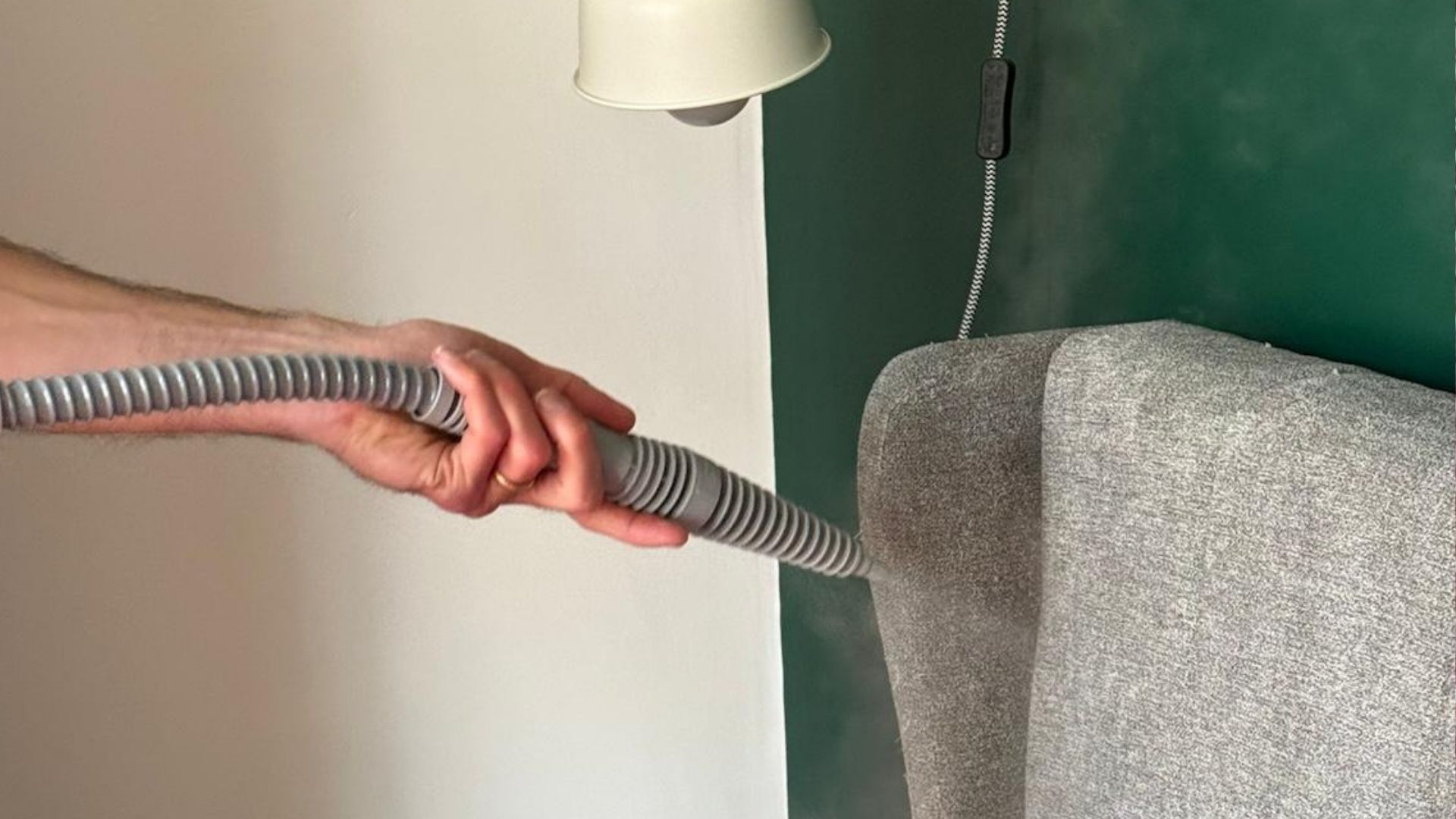 The image shows a man directing the head of a steam cleaner towards a grey fabric headboard, which is placed against a green wall and underneath a cream-colored wall light