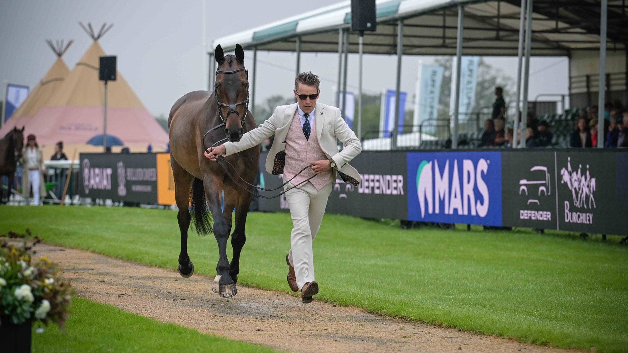 Simon Grieve, wearing a pale suit and pink waistcoat, trots up bay horse Autograf at Burghley 2026