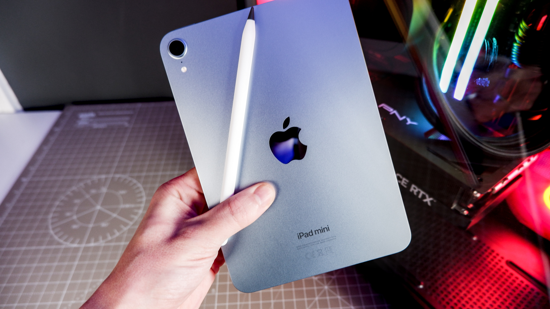 An iPad Mini A17 and iPhone 16 on a desk with a gaming PC in the background.