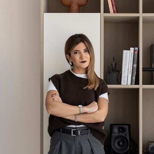 Image of a woman in gray trousers, a white T-shirt, and a brown sweater vest standing in front of a wooden, minimalist built-in.