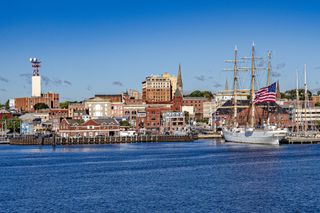 The image shows a view of the downtown of New London, Connecticut from a boat in the Long Island Sound with the Coast Guard cutter Eagle, a sailing ship, docked in the port.