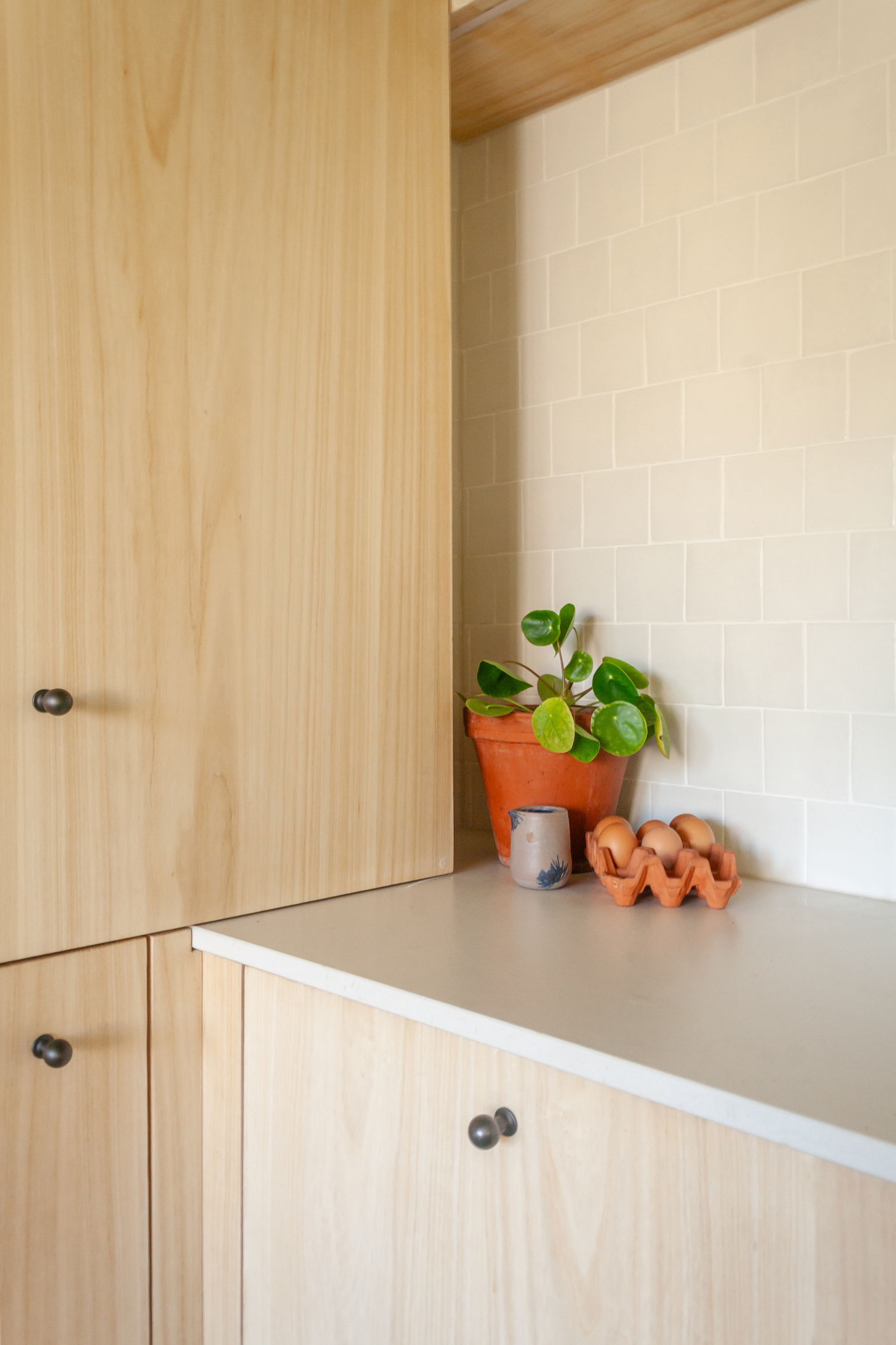 A view of the light wood cupboards in the corner of the kitchen, with the white tiled back splash and a plant pot, and some eggs in the corner.