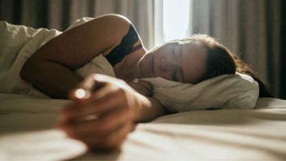Woman learning how to sleep in the heat with curtains open behind her, arm outstretched towards the camera