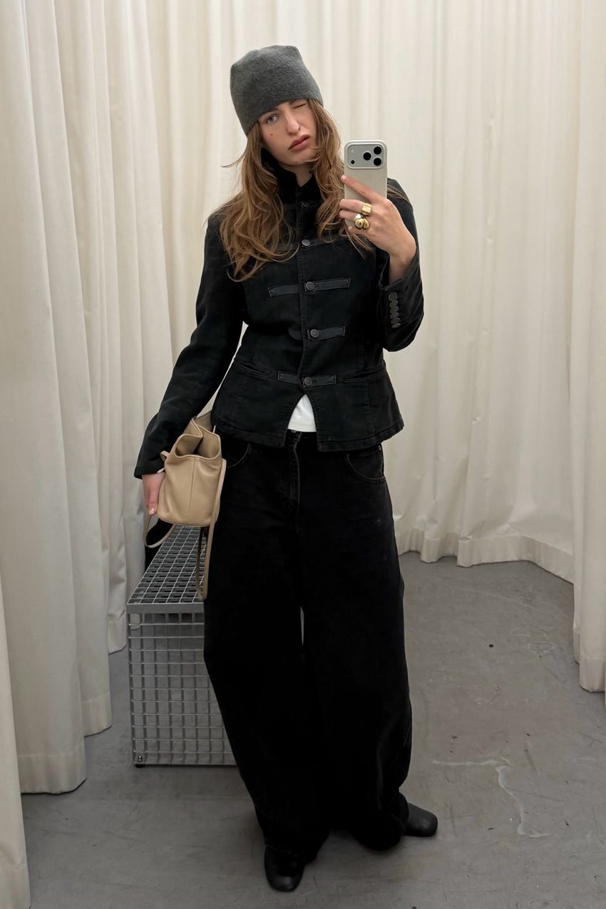 A woman standing inside a dressing room taking a selfie wearing a black military jacket over a white t-shirt, baggy black pants, black flat square-toe boots, a tan handbag, and a gray beanie hat.