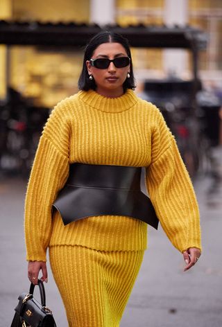 One of the best designer belts is shown in a close street-style photo of a woman wearing black sunglasses with a yellow sweater and matching knit skirt styled with a loewe obi belt and a black Hermes bag