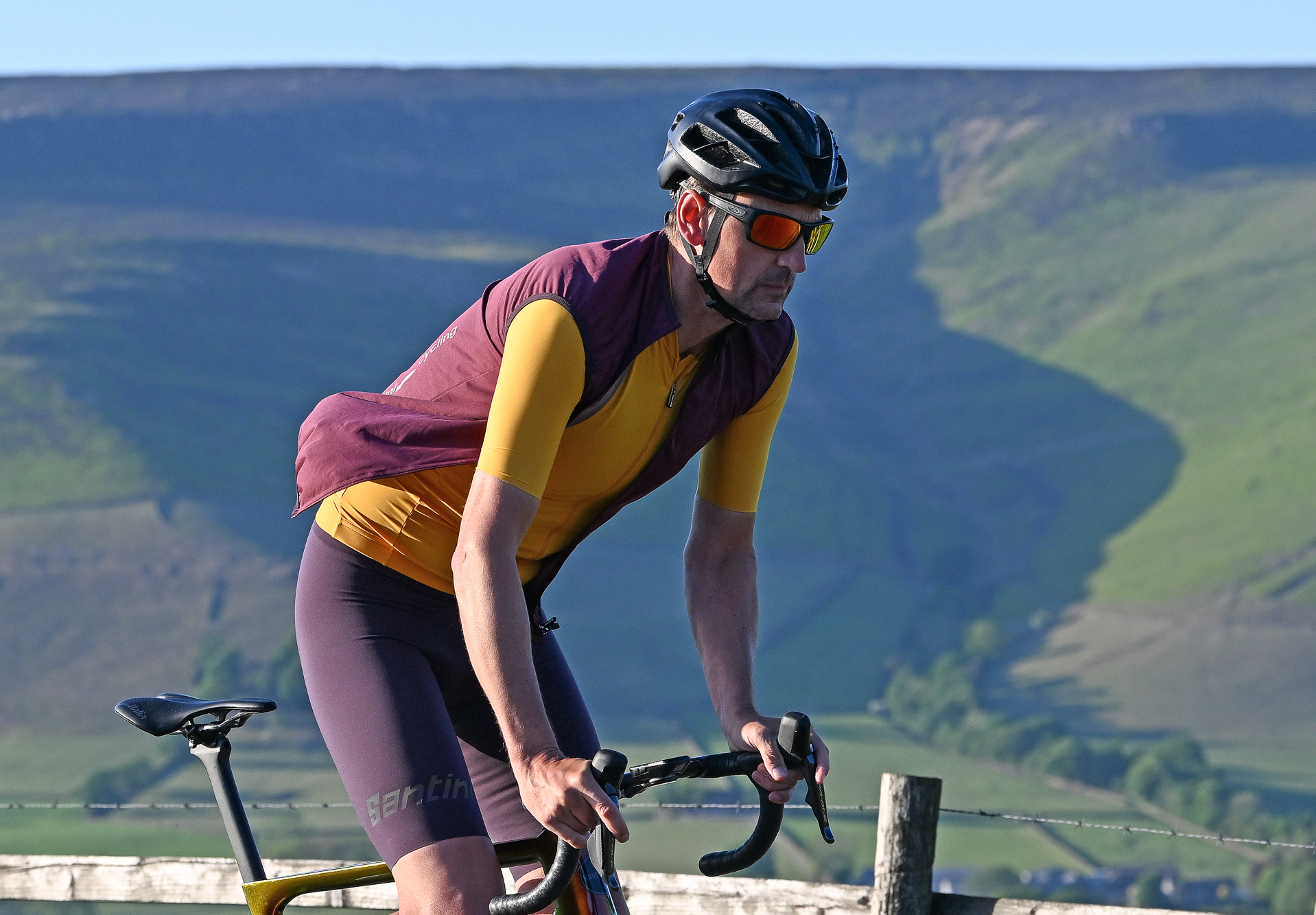 Man wearing a black helmet, sunglasses, yellow jersey and an unzipped purple gilet riding up a hill