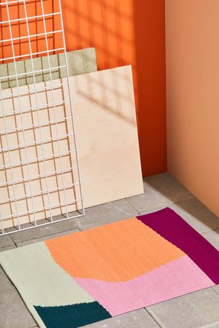 Colourful bath mat placed on a square concreate floor with orange walls. A caged frame and wood sheets resting on the wall