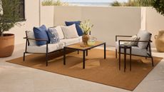 image of a tan, jute-effect outdoor rug in a cream, stone porch area. There is a white sofa, a matching accent chair, and a small wooden coffee table.