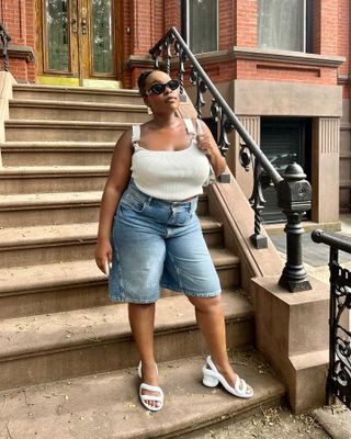 aniyah poses on stoop in brookly wearing a white tank top tucked into long denim shorts and white sandals