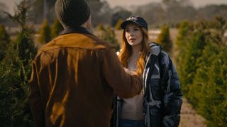 Ian Harding in a brown jacket with his back turned to the camera, arguing with Lindsey Lohan in a puffer jacket and baseball cap in the middle of a Christmas Tree farm