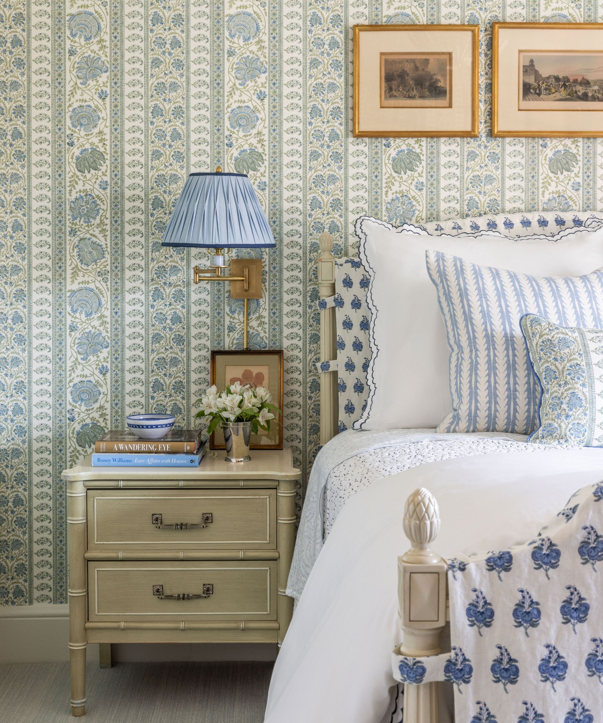 Bedroom with paisley striped green and blue wallpaper, a wooden white bed with fabric upholstery, white bed linen with blue floral cushions, and a bamboo nightstand with books and a sconce above it.