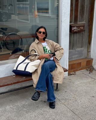 Woman wears a trench coat, bootcut jeans, graphic t-shirt, and black leather Birkenstock Boston clogs.