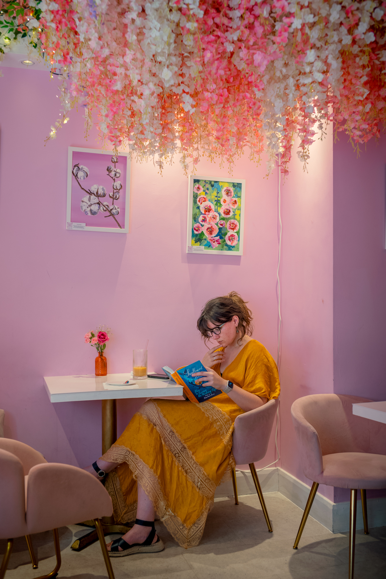A woman in a mustard-yellow dress sits reading at a small table in a pink caf&amp;eacute;, beneath a canopy of hanging pink and white artificial blossom.