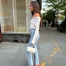 Jordanna Sharp wearing a white off-the-shoulder top, high-waisted jeans, red heels, and a white The Row bag.