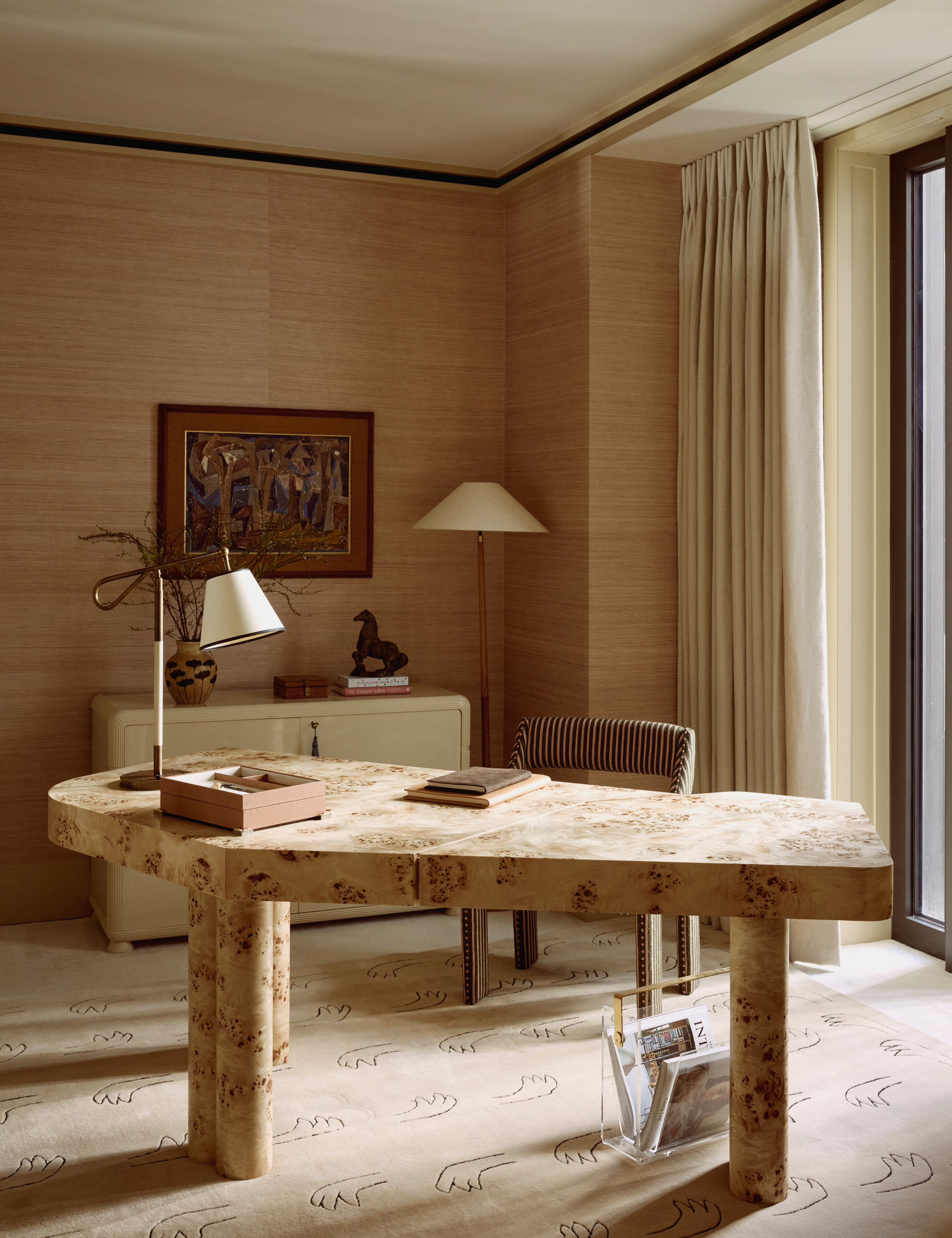 Office space featuring an asymmetrical burl wood desk, glossy file cabinet, figural motif rug, striped desk chair, and linen-shaded lamps, which rest on the desktop and floor.