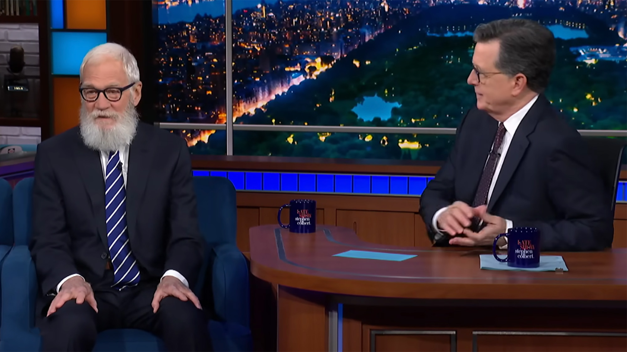 David Letterman sits on a chair and looks at the camera while Stephen Colbert interviews him from his Late Show desk. 