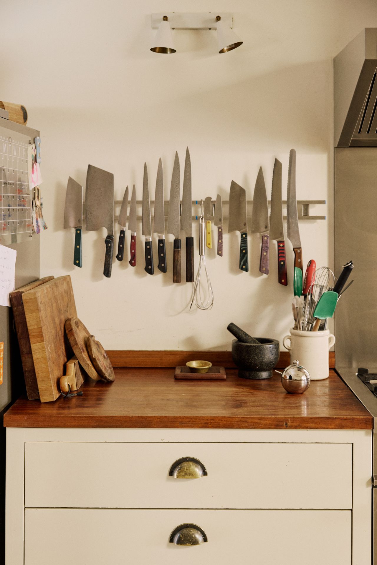 Image of a section of wooden countertop in a white kitchen with multiple wooden cutting boards, a jar of utensils, and a metal knife holder on the wall.