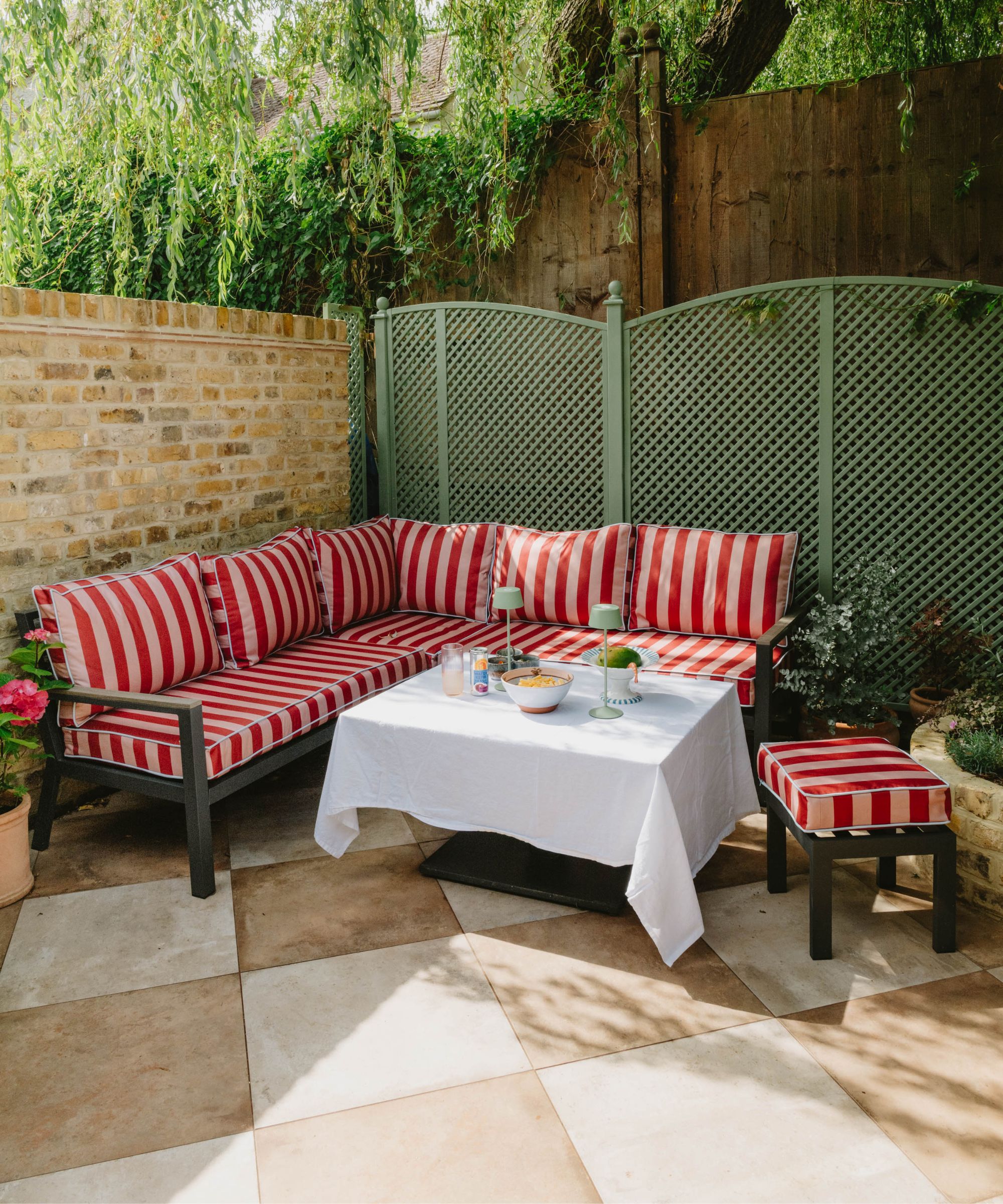 corner backyard patio with a brick wall, green painted trellis fence, beige and white checkerboard patio tiles, with a large corner sofa with pink striped cushions