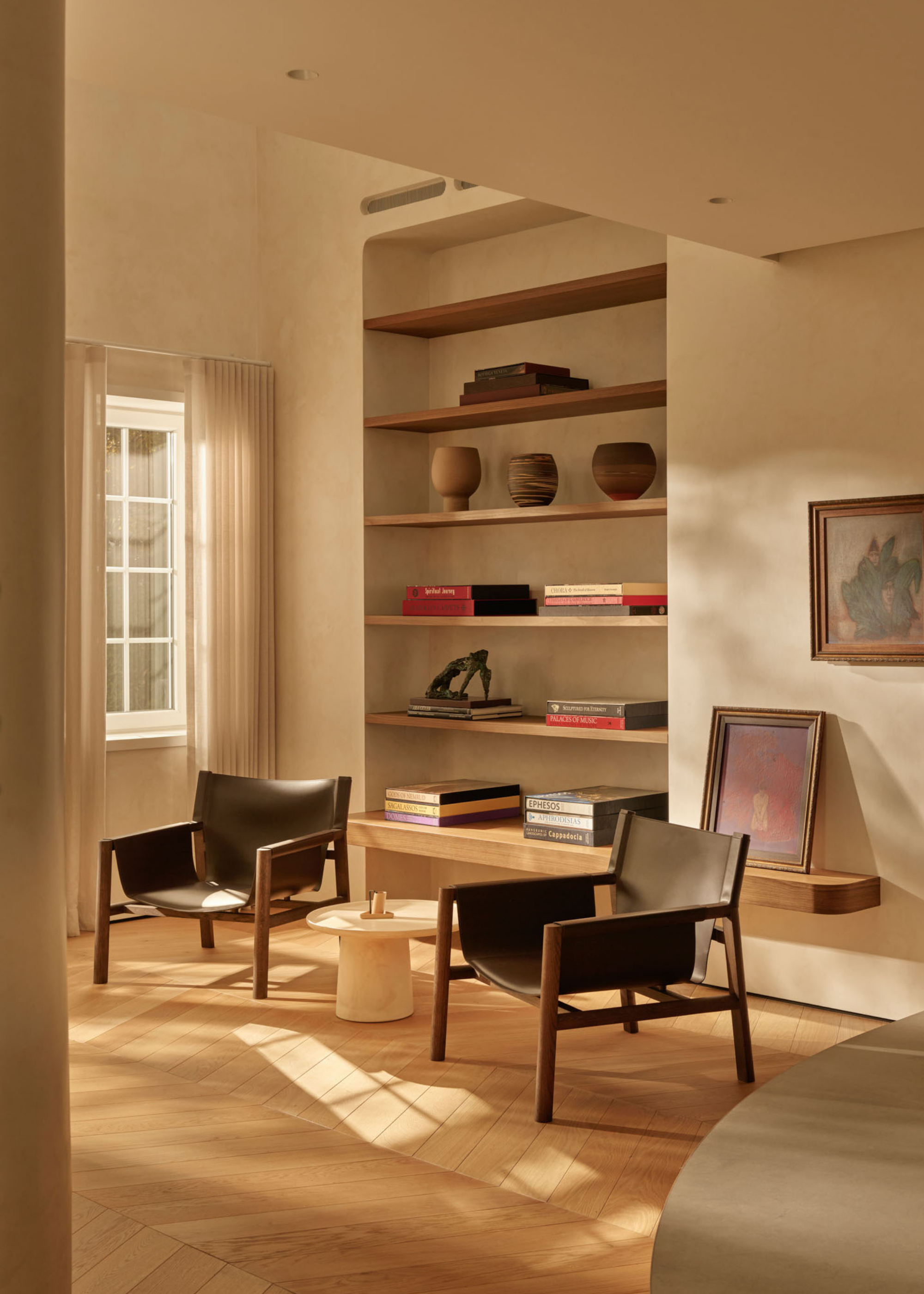 A beige living room corner iwth a shelf of books, and ceramics, by a pair of wood and leather armchairs beside a marble side table