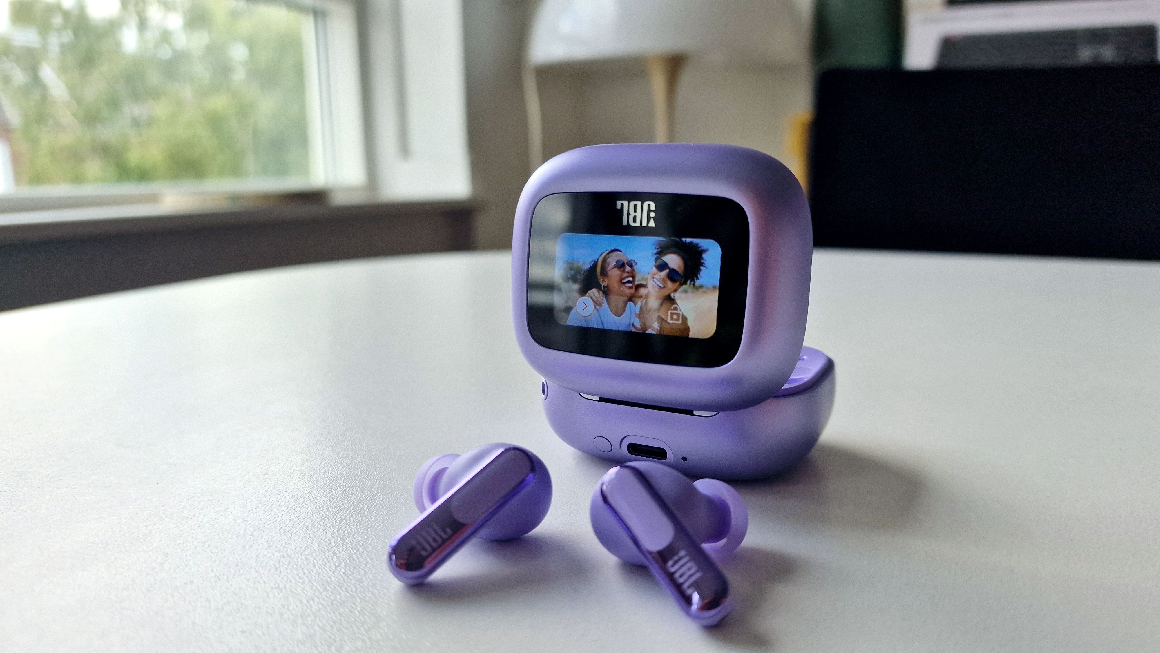 The JBL Live Beam 3 truly wireless earphones in purple, photographed on a table next to their charging case.