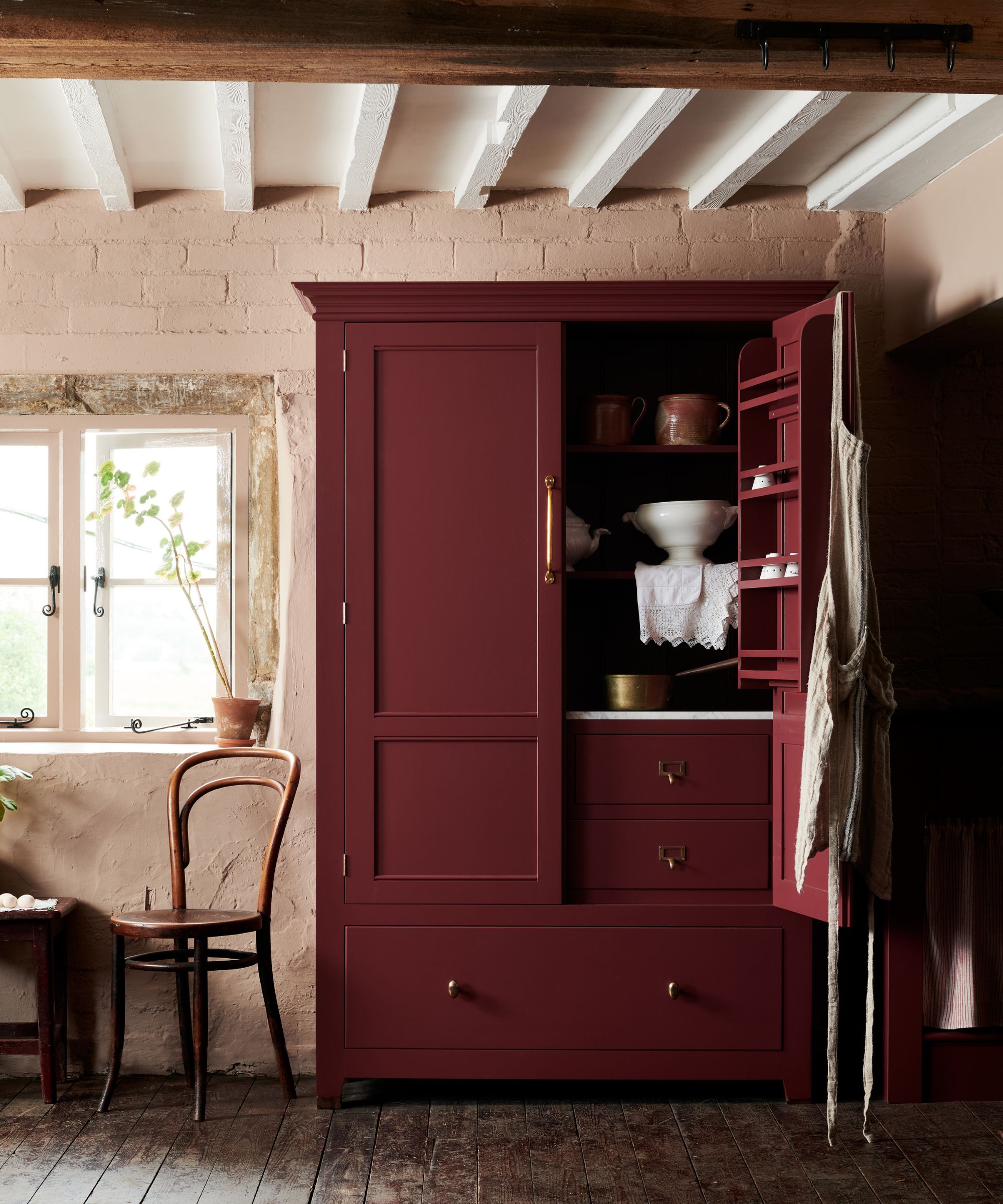 Burgundy-red pantry cabinet in deVOL's Refectory Red, set against rustic plaster walls, exposed timber beams, and aged wood flooring.