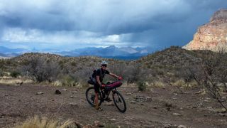 Sarah Sturm is riding her loaded bike along a gravel path with a dramatic cloudy sky in the background