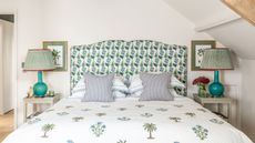 A bright white attic bedroom space with a large double bed with a blue and green floral headboard. The bedding is white with botanical print, and there are two striped cushions on the bed. To either side of the bed are white bedside tables with blue lamps and striped lampshades, and above each are matching botanical prints in gold frames with green backing.