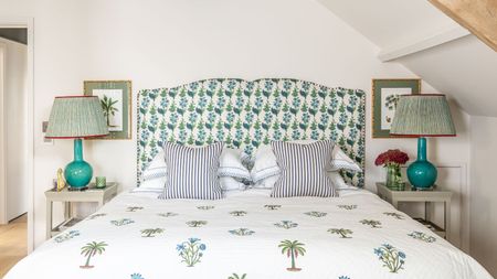 A bright white attic bedroom space with a large double bed with a blue and green floral headboard. The bedding is white with botanical print, and there are two striped cushions on the bed. To either side of the bed are white bedside tables with blue lamps and striped lampshades, and above each are matching botanical prints in gold frames with green backing.