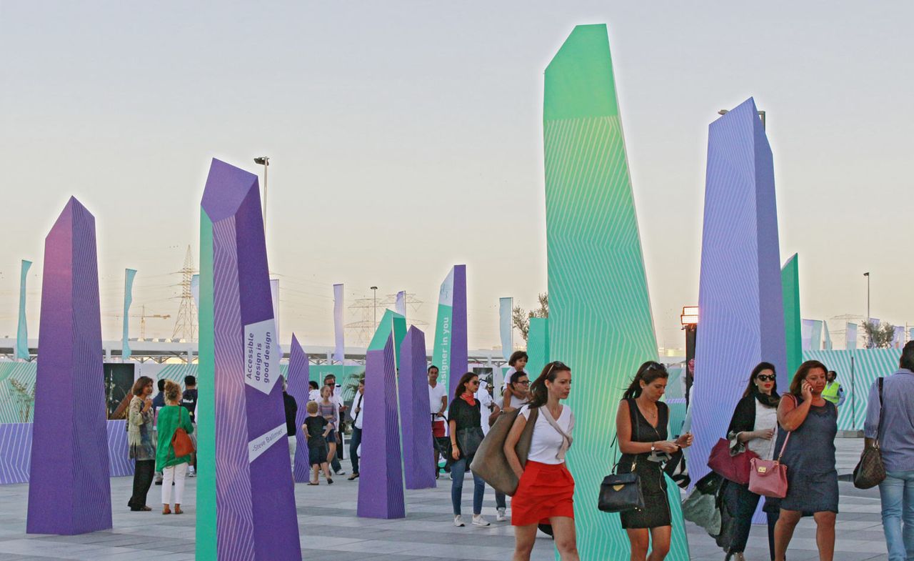 The entrance space at the Dubai Design District. People are walking between the asymmetrical &#039;stalagmite&#039; looking decorative installations in green and purple hues.