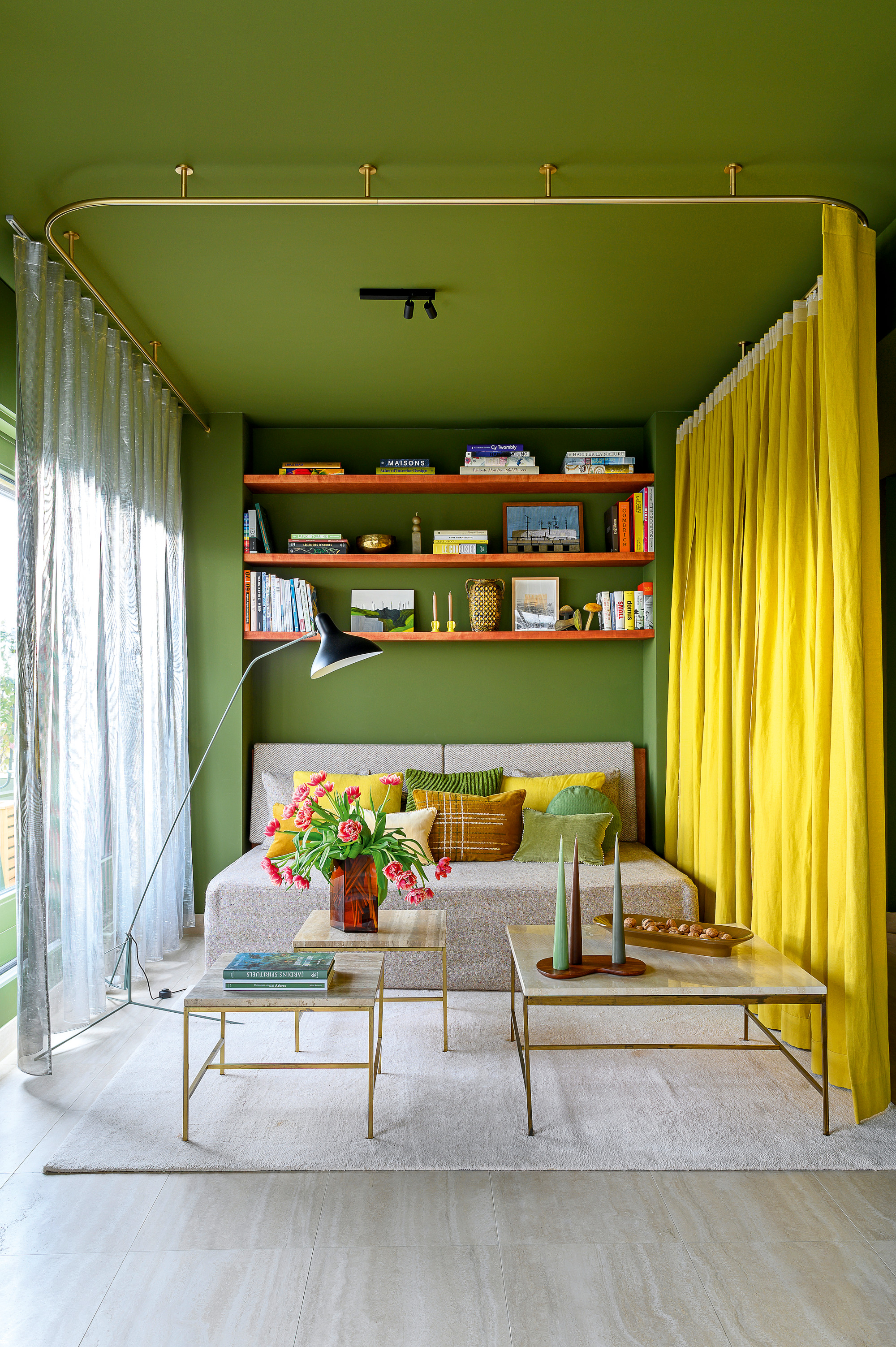 Living area with matcha green walls, grey sofa bed, marble and brass coffee tables, black and chrome floor lamp and yellow curtain to create privacy