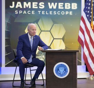 a man in a blue suit sits on a chair next to a podium. a backdrop shows hexagonal shapes arranged together below the words 'james webb space telescope' a flag is draped on a pole on the right.