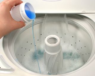 A cup of blue laundry detergent being poured into a top-loading washing machine