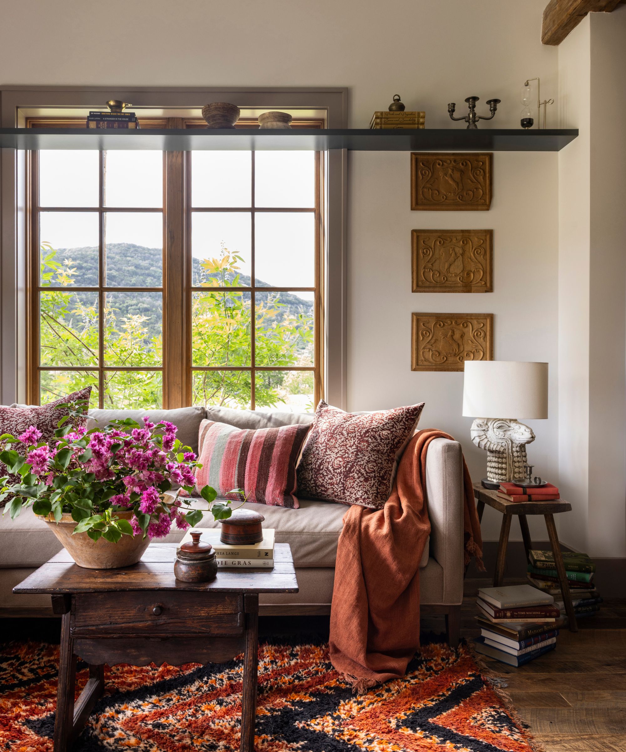 A cozy living room with neutral walls, large windows, a taupe sofa with warm pink cushions, a red patterned rug, and a wooden coffee table with a vase of pink flowers.