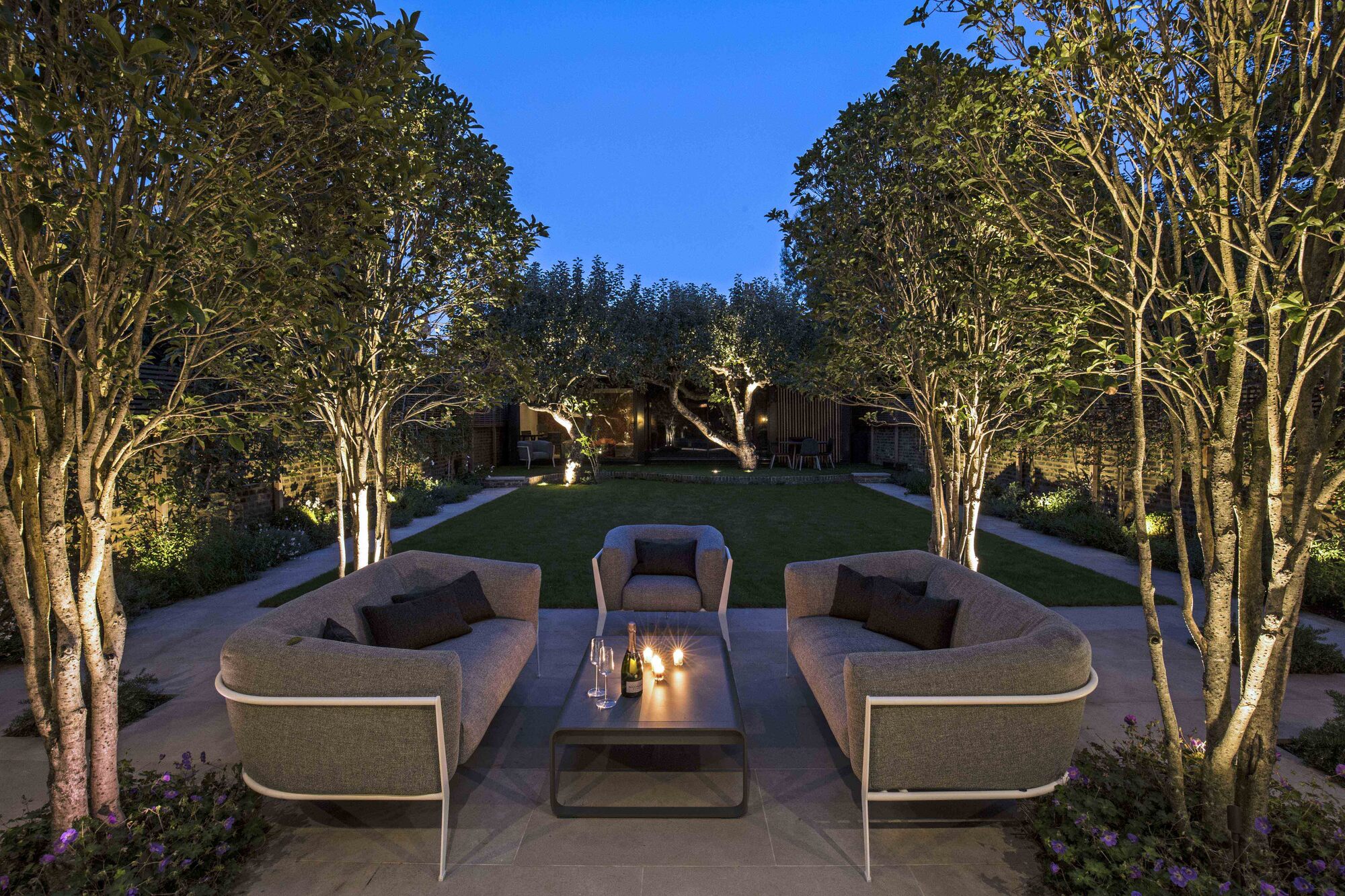 A garden at dusk with two sofas on a patio, candles on a table, and trees with uplighting lining the lawn