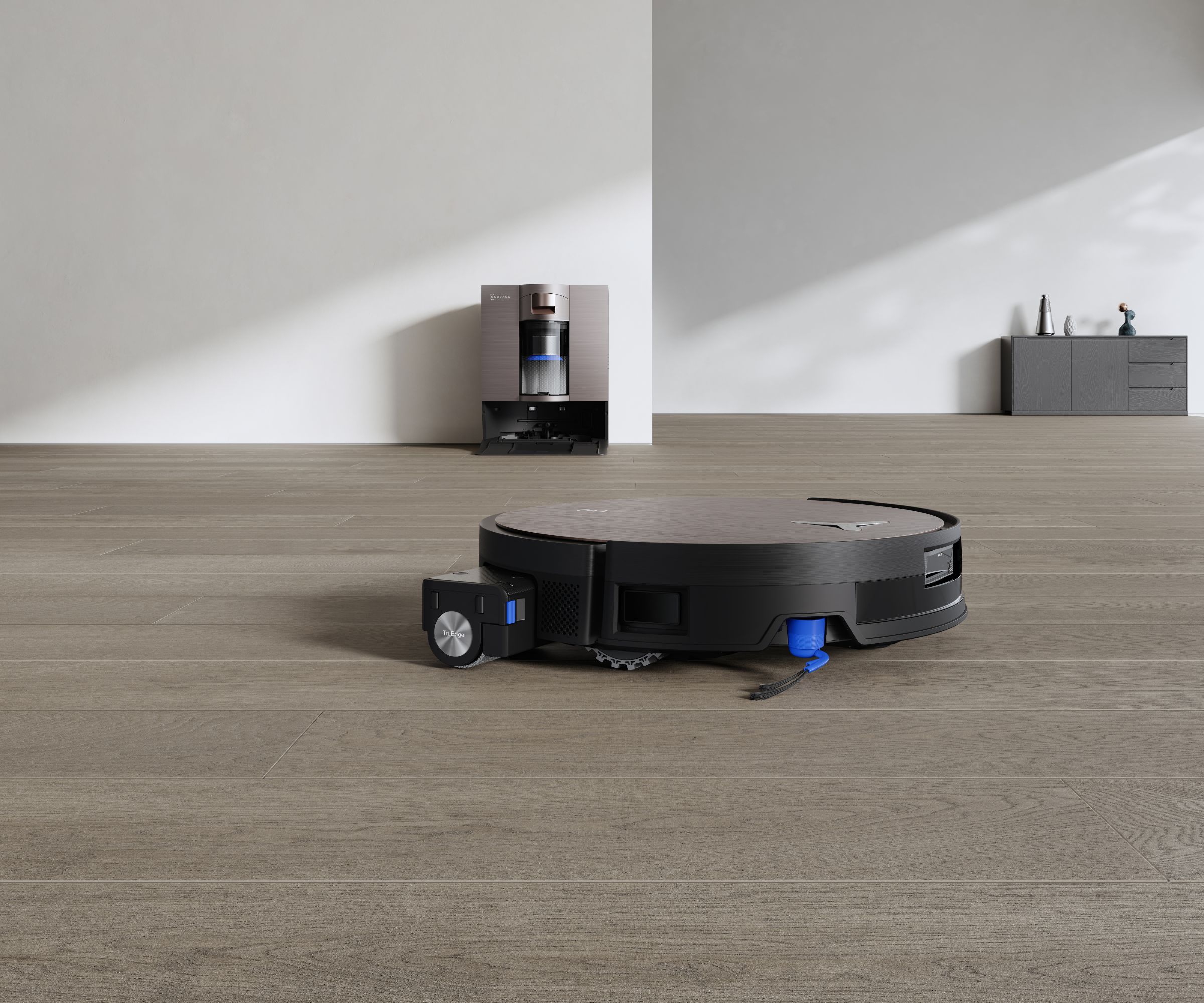 ECOVACS DEEBOT X11 OMNICYCLONE on brown floor, pictured in front of silver cleaning station and white walls.