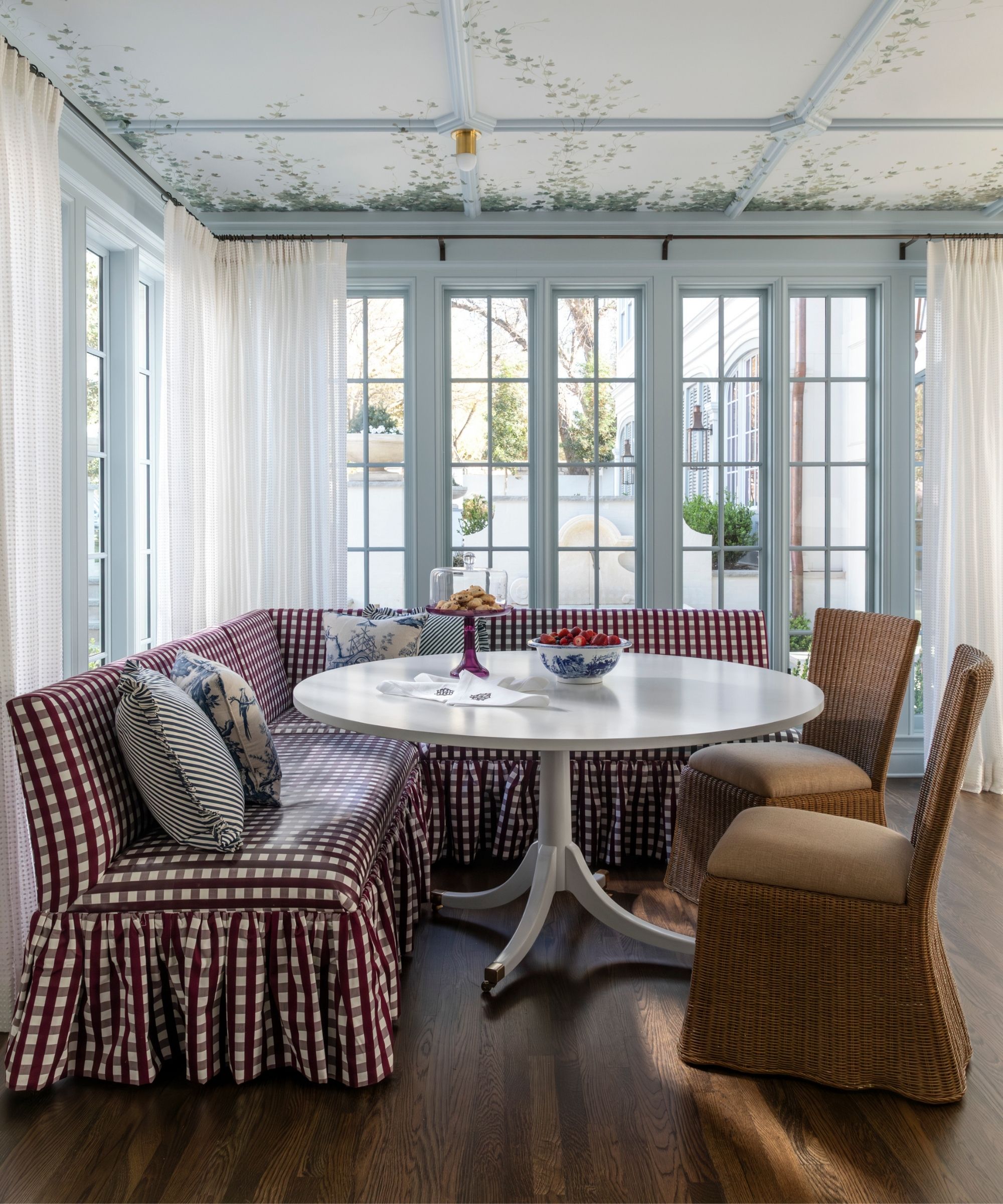 Breakfast nook with pale blue painted windows and moulding on ceiling, a mural ceiling, sheer drapes, a red gingham skirted bench, two rattan chairs, and a round white breakfast table.