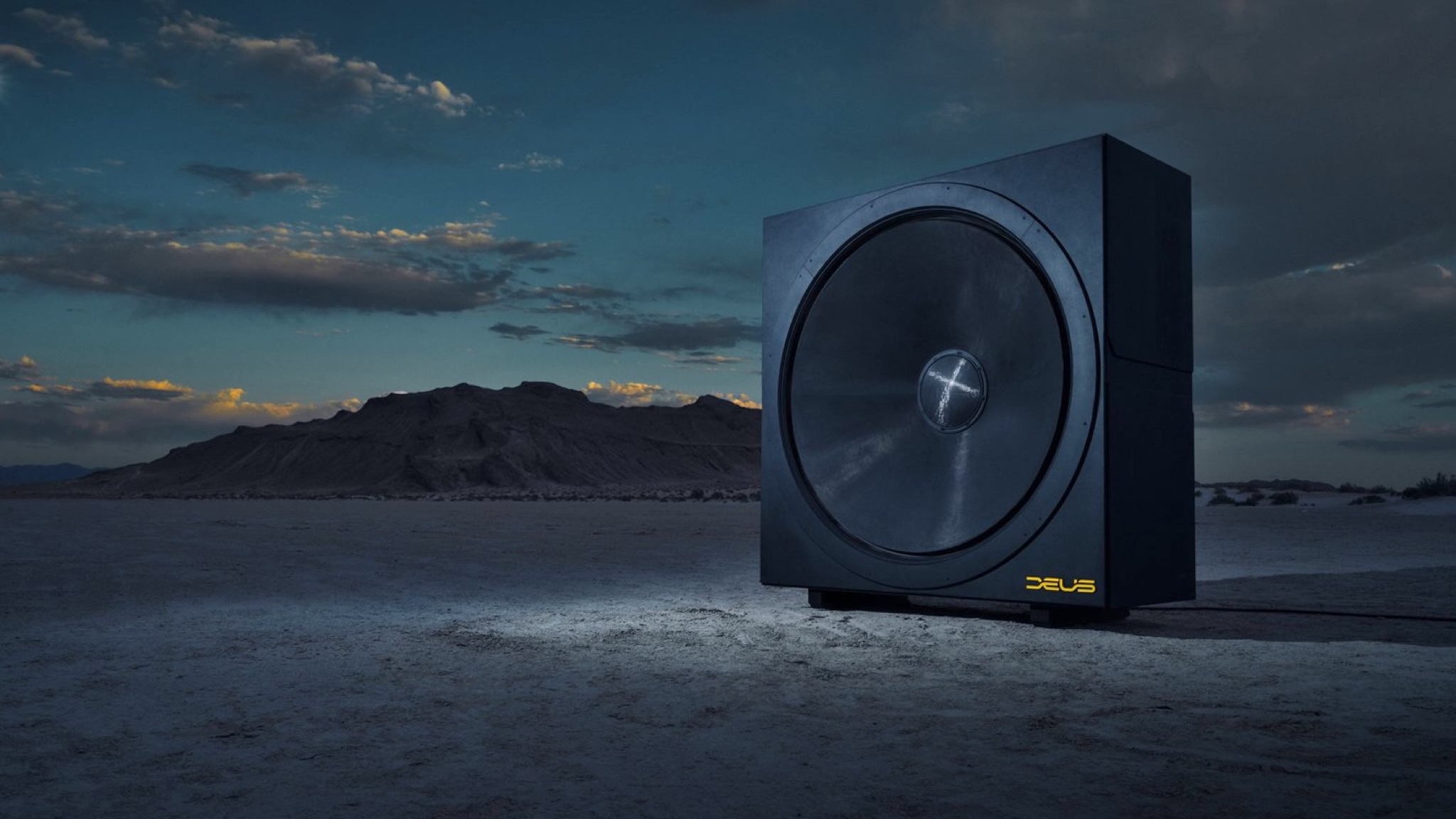 A promotional photo of the Deus Invicta 60 subwoofer on a dried lake bed underneath a late evening sky