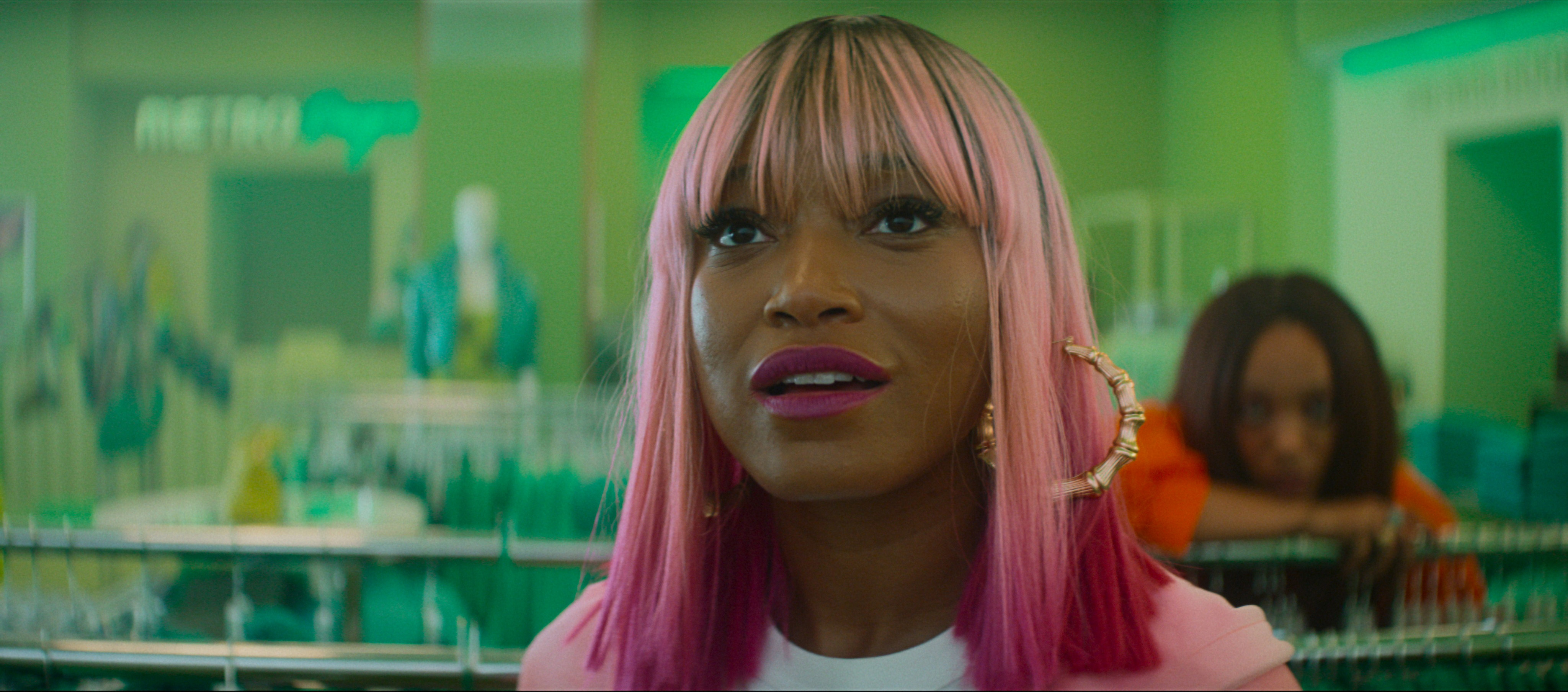 A woman wearing a pink bob wig (Keke Palmer as Corvette) stands in an all-green clothing store as a second blurred woman (Naomi Ackie as Sade) leans on a rack behind her, in 'I Love Boosters'