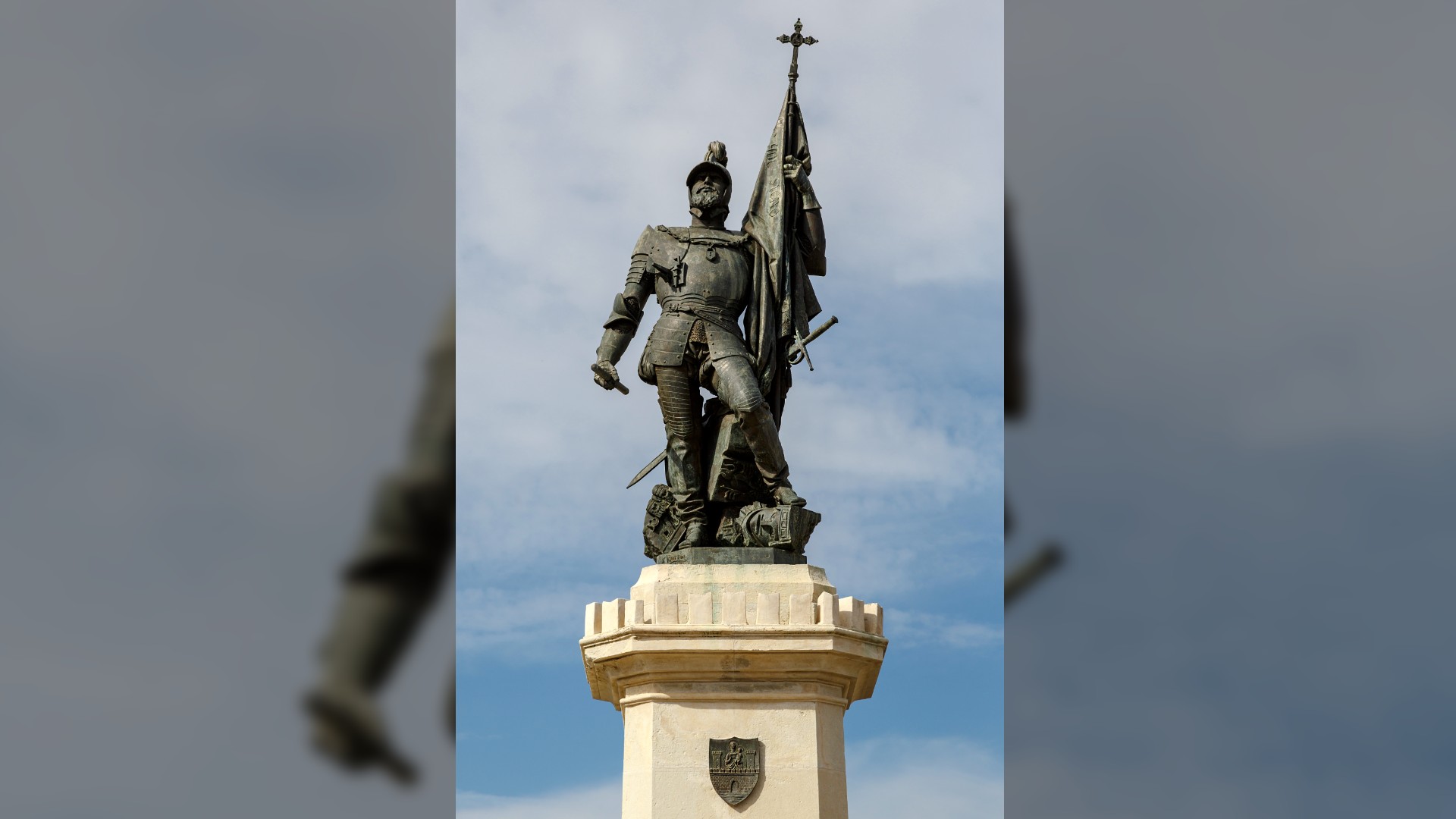 Statue of Hernan Cortes in Medellin, Spain. Here we see him standing tall wearing armor and a helmet (complete with a plume on top). He’s holding a flag/banner with a small cross on the top. He is standing upon a plinth which looks like a castle tower. On the front of the plinth is a small shield which depicts a knight standing upon a castle rampart.