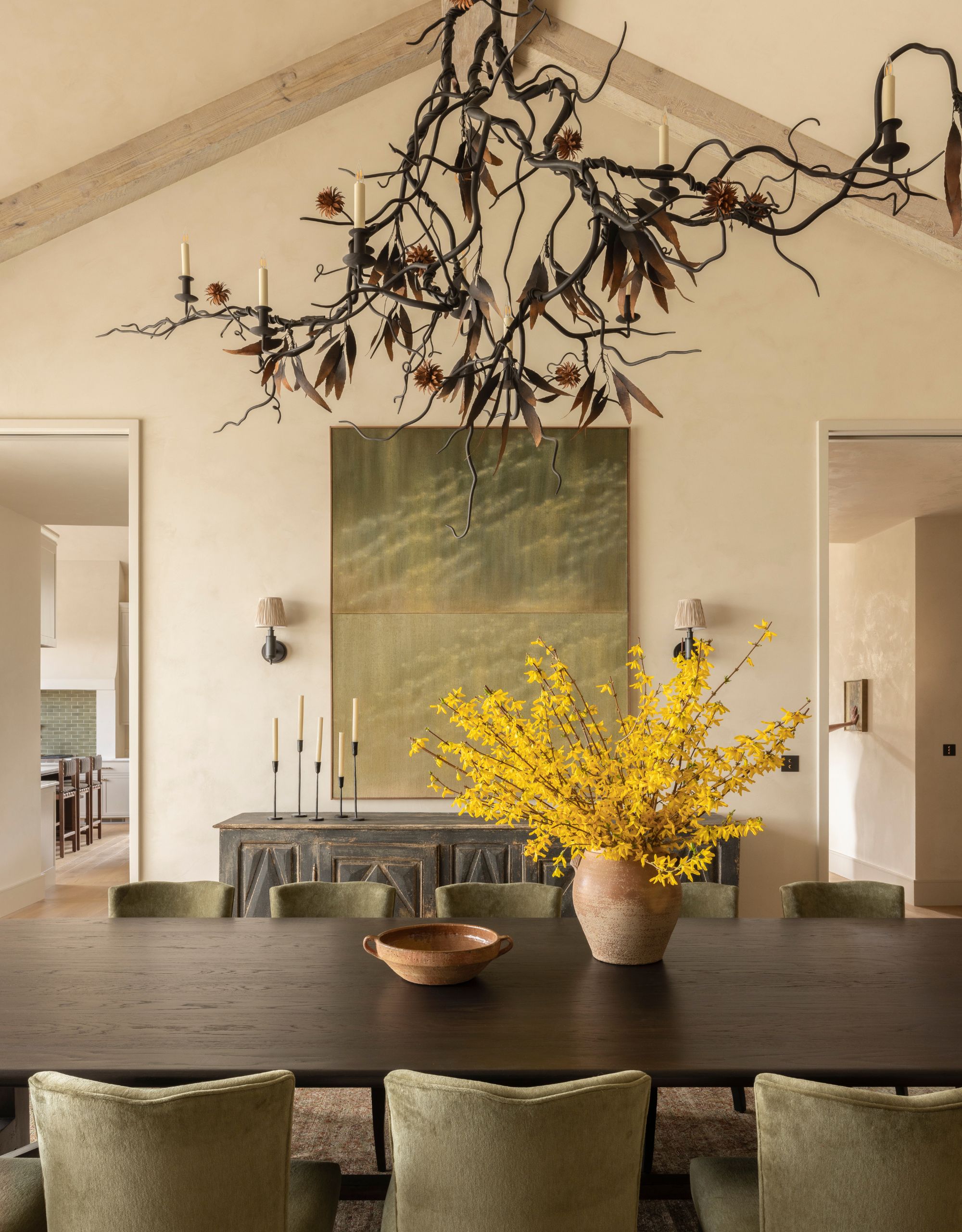 A dining area with a large dark wood dining table with green velvet dining chairs, in front of a large green artwork on the white walls with a foliage-inspired chandelier with candles