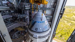 A white and blue Boeing Starliner capsule is hoisted atop its rocket in a huge hangar.