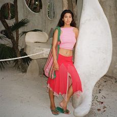 michelle li leands on building pillar while wearing a pink tank top and a red midi skirt and green flip flops and she is carrying a large crochet shoulder bag